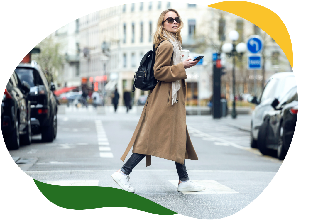 Šiuolaikiška moteris su dideliais akiniais nuo saulės ir ilgu smėlio spalvos paltu eina per gatvę, dešinėje rankoje laikydama mobilųjį telefoną