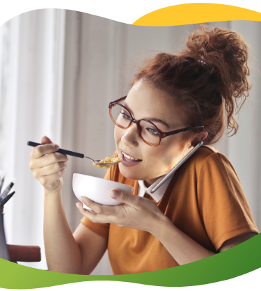 Jauna moteris su akiniais ir viršugalvyje surištais plaukais sėdi priešais nešiojamąjį kompiuterį, kalba telefonu ir valgo